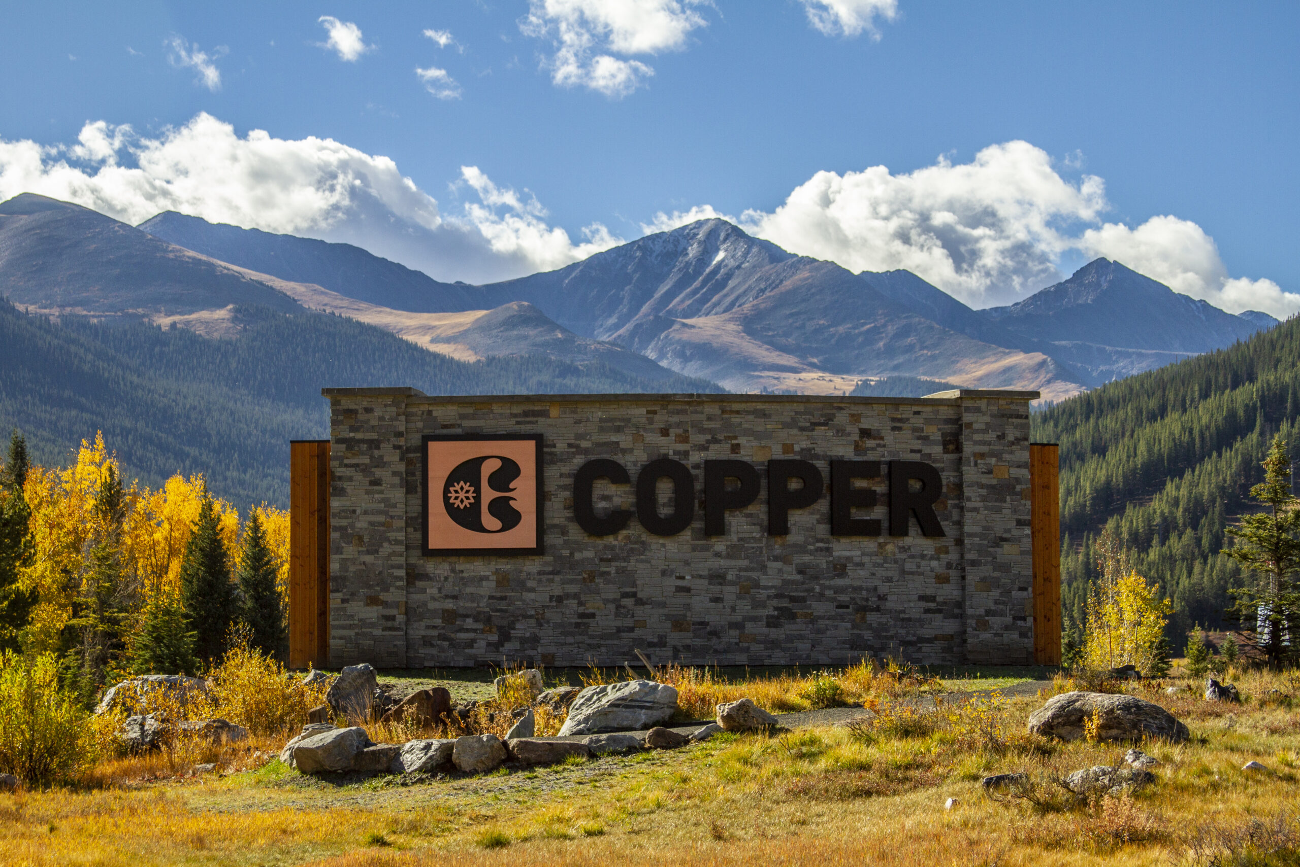 Sign with "Copper" and a logo on a stone structure, set against a mountainous landscape with trees and a clear sky.